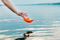 The girl lets down her paper boat to the water with her hand. An orange ship is hanging over the river