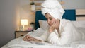 Girl reading book. Woman in bedroom with towel on her head and in white coat lies on stomach in bed and turns over page of book. Royalty Free Stock Photo
