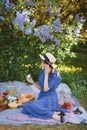 Girl in a lilac dress and a straw hat sits on a blanket in the garden and holds a delicious marshmallow in her hand