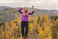 A girl in a lilac jacket makes a salfi on a mountain, a view of the mountains and an autumnal forest by a cloudy day Royalty Free Stock Photo