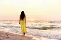 A girl in a long dress with long black hair walks at dawn along a deserted beach along