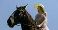 Girl in long dress sitting astride the dark horse by holding its reins. They are outdoors on a summer day, against a