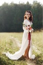 Girl in a long dress stands in a field with wreath on her head and bouquet of flowers in her hands, beautiful woman in the rays of Royalty Free Stock Photo