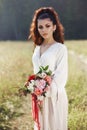 Girl in a long dress stands in a field with wreath on her head and bouquet of flowers in her hands, beautiful woman in the rays of Royalty Free Stock Photo