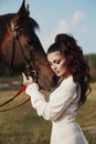 Girl in a long dress stands near a horse, a beautiful woman strokes a horse and holds the bridle in a field in autumn. Country Royalty Free Stock Photo