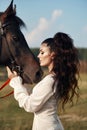 Girl in a long dress stands near a horse, a beautiful woman strokes a horse and holds the bridle in a field in autumn. Country Royalty Free Stock Photo