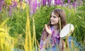 Girl with long hair sits in a clearing with lupine flowers and plays with a tambourine