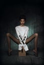 A girl with long legs in black tights and a white straitjacket sits on a wooden pedestal with her legs spread to the side Royalty Free Stock Photo