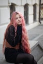 Girl with long pink hair sits on the steps and looks ahead, is wearing a Basco belt