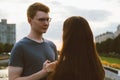 Girl with long thick dark hair holding hands redhead boy in blue t-shirt on bridge, teen love at evening. Boy looks tenderly at