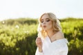 Girl in a long white dress with a dandelion flower in her hand stands in grass in a field. Blonde woman in the sun in a light Royalty Free Stock Photo