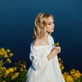 Girl in a long white dress with a flower in her hand stands near the lake. Blonde woman in the sun in a light dress. Girl resting Royalty Free Stock Photo