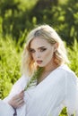 Girl in a long white dress sits on the grass in a field. Blonde woman in the sun in a light dress. Girl resting and dreaming, Royalty Free Stock Photo