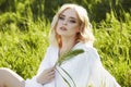 Girl in a long white dress sits on the grass in a field. Blonde woman in the sun in a light dress. Girl resting and dreaming, Royalty Free Stock Photo