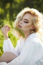 Girl in a long white dress sits on the grass in a field. Blonde woman in the sun in a light dress. Girl resting and dreaming, Royalty Free Stock Photo