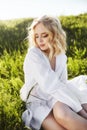 Girl in a long white dress sits on the grass in a field. Blonde woman in the sun in a light dress. Girl resting and dreaming, Royalty Free Stock Photo