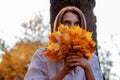 Girl are looking upwards holding bouquet of dry yellow leaves in front of face sitting on the ground under a tree in autumn park Royalty Free Stock Photo