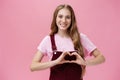 Girl loves family. Kind charming young woman in overalls with small tattoo on arm showing heart gesture over body and