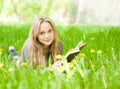 Girl lying on grass with dandelions reading a book and looking at camera Royalty Free Stock Photo