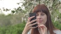 Girl makes selfie in the garden. An attractive red-haired woman smiles making selfi using a mobile phone in a cherry Royalty Free Stock Photo