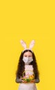 A girl with a medical mask and Easter bunny ears is holding colorful Easter eggs in a basket on a yellow background. Easter, Royalty Free Stock Photo