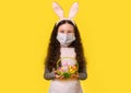 A girl with a medical mask and Easter bunny ears is holding colorful Easter eggs in a basket on a yellow background. Easter, Royalty Free Stock Photo