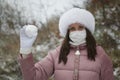 A girl in a medical mask and winter clothes on a walk in the park. Holds a ball of snow in his hand. The ground is covered with th