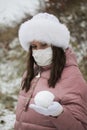 A girl in a medical mask and winter clothes on a walk in the park. Holds a ball of snow in his hand. The ground is covered with th