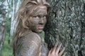 Girl with a mask of mud near a tree trunk in an ecological resort. Beautiful blonde