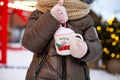 Girl with mug with snow, candy cane and inscription Merry and Bright in her hands outdoor in warm clothes in winter at festive