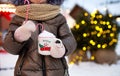 Girl with mug with snow, candy cane and inscription Merry and Bright in her hands outdoor in warm clothes in winter at festive