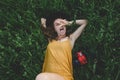 A girl in a mustard-colored dress and a hat lies on a green field next to a red camera and shows her tongue