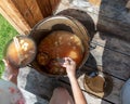 Girl in nature puts in a bowl of soup borscht with meat and bone and vegetables from a large pot on a wooden floor.