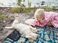 Girl petting small white dog lying on beach shore near water lake river. Friendship of domestic animal and child kid human. Royalty Free Stock Photo