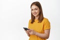 Girl with phone. Young blond woman using cellphone, texting message, smiling and looking happy at camera, standing in Royalty Free Stock Photo