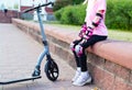 A girl in pink protective gear sits on a curb next to a scooter. The concept of safety while riding a scooter. Copy Royalty Free Stock Photo