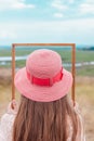 Girl in pink straw hat holds empty wooden picture frame in hands on nature landscape background faceless. Creative idea Royalty Free Stock Photo