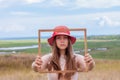 Girl in pink straw hat holds empty wooden picture frame in hands on nature landscape background portrait. Creative idea Royalty Free Stock Photo