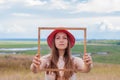 Girl in pink straw hat holds empty wooden picture frame in hands on nature landscape background portrait. Creative idea Royalty Free Stock Photo