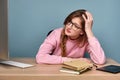 A girl in a pink sweater sits at a table with a computer and books, and, holding on to her head, looks exhaustedly at