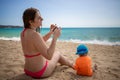 Girl in a pink swimsuit sits on beach and photographs with smartphone her little cute baby next to her in an orange T
