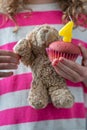 A girl in a pink and white shirt holding a teddy bear waving and a pink cupcake with a candle with the number four, close-up