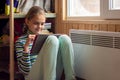 A girl plays in a tablet sitting next to a radiator by a window in a country house