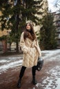 Girl posing on road on winter background. Glamorous funny young woman with smile wearing stylish creamy long fur coat