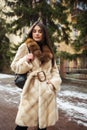 Girl posing on road on winter background. Glamorous funny young woman with smile wearing stylish creamy long fur coat