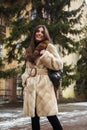 Girl posing on road on winter background. Glamorous funny young woman with smile wearing stylish creamy long fur coat