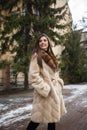 Girl posing on road on winter background. Glamorous funny young woman with smile wearing stylish creamy long fur coat