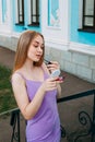 Girl in a purple dress paints her lips with lipstick, near the blue facade of a beautiful old building