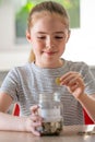 Girl Putting Coins Into Glass Jar Labelled Savings At Home