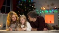 Girl putting letter for Santa into envelope, lying under X-mas tree with parents